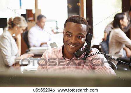 Stock Photography - Businessman On Phone At Desk With Meeting In Background. Fotosearch