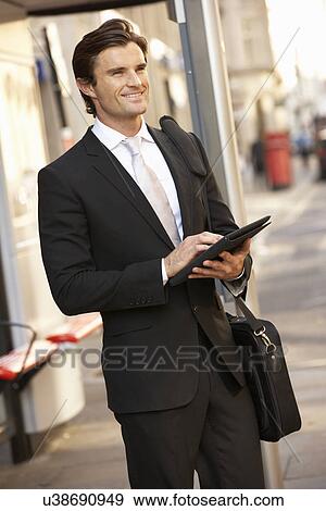 Stock Photo - Businessman standing at bus stop using tablet. Fotosearch