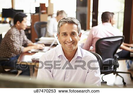 Stock Image - Businessman Working At Desk With Meeting In Background. Fotosearch
