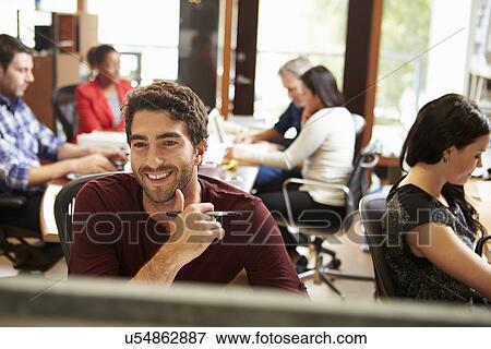 Stock Photo - Businessman Working At Desk With Meeting In Background. Fotosearch