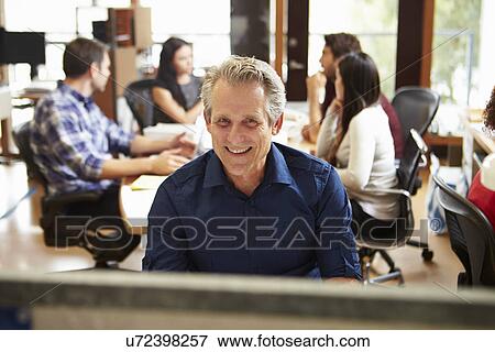 Stock Photo - Businessman Working At Desk With Meeting In Background. Fotosearch