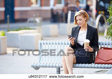 Stock Photo - Businesswoman On Park Bench With Coffee Using Mobile Phone. Fotosearch