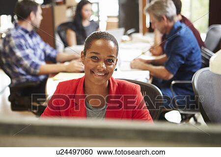 Stock Image - Businesswoman Working At Desk With Meeting In Background. Fotosearch