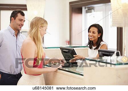Stock Image - Couple Checking In At Hotel Reception Using Digital Tablet. Fotosearch