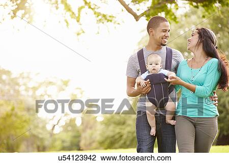 Stock Image - Family With Baby Son In Carrier Walking Through Park. Fotosearch