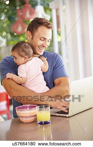 Stock Image - Father Holding Baby And Making Snack Whilst Using Laptop. Fotosearch