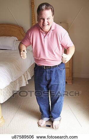 Stock Photograph - Happy Overweight Man Standing On Scales In Bedroom. Fotosearch