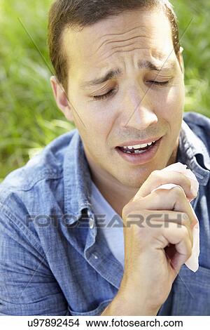 Picture - Man sitting on grass sneezing. Fotosearch