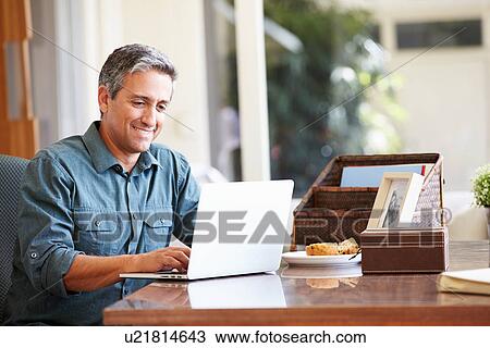 Stock Image - Mature Hispanic Man Using Laptop On Desk At Home. Fotosearch