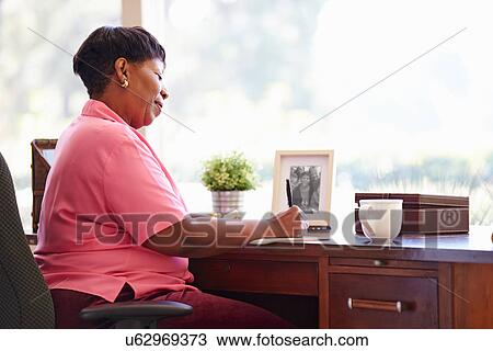 Stock Image - Mature Woman Writing In Notebook Sitting At Desk. Fotosearch
