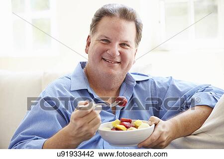 Overweight Man Sitting On Sofa Eating Bowl Of Fresh Fruit View Large Photo Image Stock Image - Overweight Man Sitting On Sofa Eating Bowl Of Fresh Fruit. Fotosearch