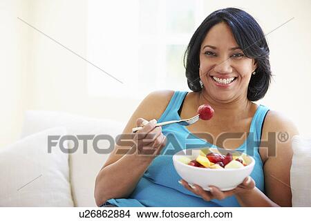 Picture - Overweight Woman Sitting On Sofa Eating Bowl Of Fresh Fruit. Fotosearch