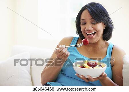 Stock Photo - Overweight Woman Sitting On Sofa Eating Bowl Of Fresh Fruit. Fotosearch