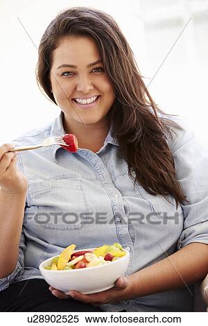 Stock Photography - Overweight Woman Sitting On Sofa Eating Bowl Of Fresh Fruit. Fotosearch