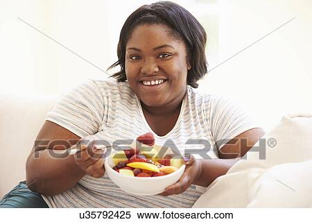 Stock Photography - Overweight Woman Sitting On Sofa Eating Bowl Of Fresh Fruit. Fotosearch