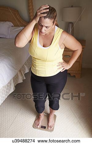 Picture - Overweight Woman Weighing Herself On Scales In Bedroom. Fotosearch