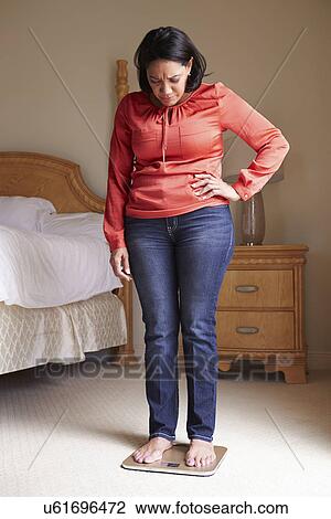 Stock Image - Overweight Woman Weighing Herself On Scales In Bedroom. Fotosearch