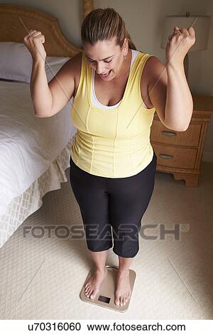 Stock Image - Overweight Woman Weighing Herself On Scales In Bedroom. Fotosearch