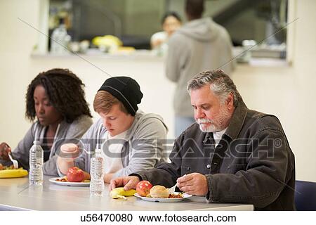 Stock Image - People Sitting At Table Eating Food In Homeless Shelter. Fotosearch