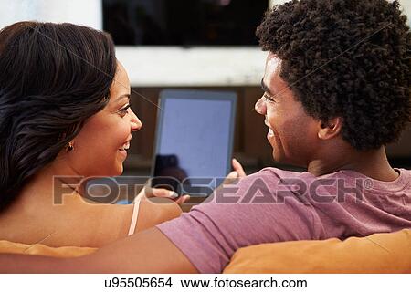 Picture - Rear View Of Couple Sitting On Sofa Using Digital Tablet. Fotosearch