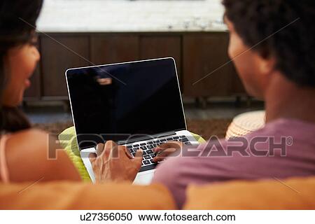 Stock Image - Rear View Of Couple Sitting On Sofa Using Laptop. Fotosearch