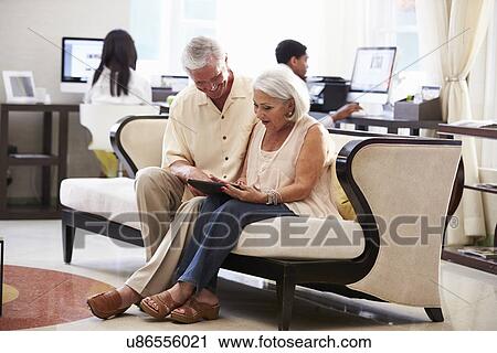 Stock Image - Senior Couple In Hotel Lobby Looking At Digital Tablet. Fotosearch