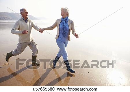 Stock Image - Senior Couple Running Along Winter Beach. Fotosearch
