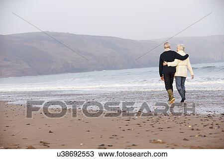 Stock Image - Senior Couple Walking Along Winter Beach. Fotosearch