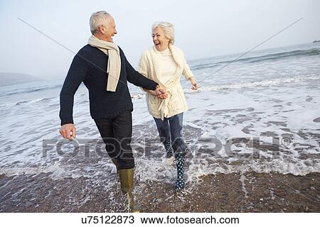 Stock Image - Senior Couple Walking Along Winter Beach. Fotosearch