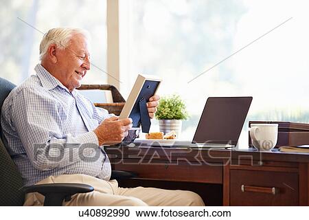 Stock Image - Senior Man Sitting At Desk Looking At Photo Frame. Fotosearch