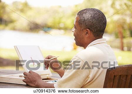 Stock Photograph - Senior Man Sitting At Outdoor Table Painting Landscape. Fotosearch