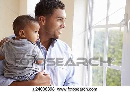 Stock Photo - Smiling Father Standing By Window With Baby Son At Home. Fotosearch