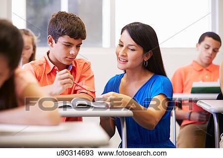 Stock Image - Teacher Helping Male High School Student In Classroom. Fotosearch