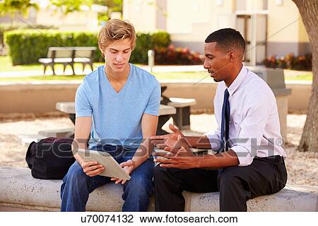 Stock Image - Teacher Sitting Outdoors Helping Male Student With Work. Fotosearch