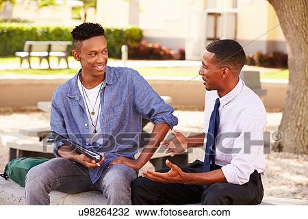 Stock Image - Teacher Sitting Outdoors Helping Male Student With Work. Fotosearch