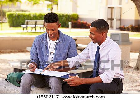 Stock Photo - Teacher Sitting Outdoors Helping Male Student With Work. Fotosearch