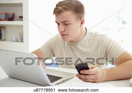 Stock Image - Teenage Boy Sending Text Message Whilst Studying On Laptop. Fotosearch