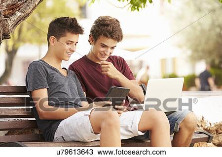Stock Image - Teenage Boys On Park Bench Using Laptop And Digital Tablet. Fotosearch