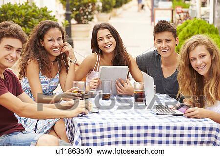 Stock Image - Teenage Friends Sitting At Cafe Using Digital Devices. Fotosearch