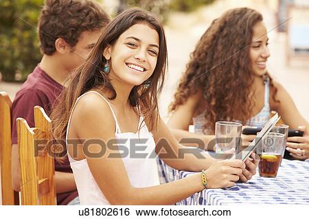Stock Photograph - Teenage Friends Sitting At Cafe Using Digital Devices. Fotosearch