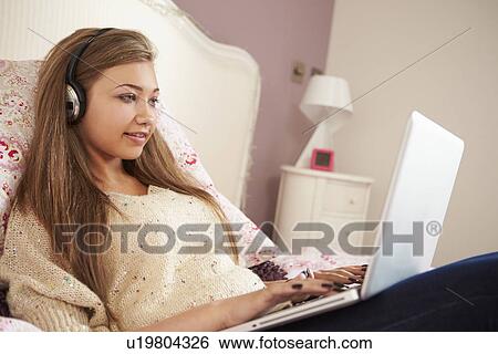 Stock Photograph - Teenage Girl Lying On Bed Using Laptop Wearing Headphones. Fotosearch