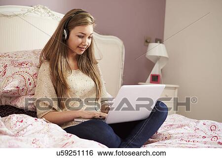 Stock Photograph - Teenage Girl Lying On Bed Using Laptop Wearing Headphones. Fotosearch