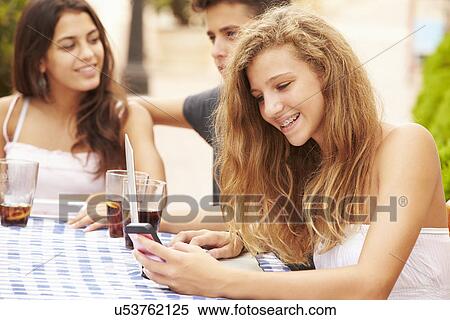 Stock Photography - Teenage Girl Using Mobile Phone Sitting At Cafe With Friends. Fotosearch