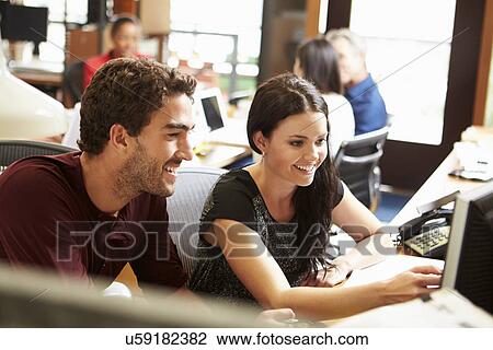 Stock Image - Two Colleagues Working At Desk With Meeting In Background. Fotosearch