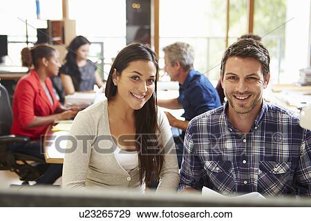 Two Colleagues Working At Desk With Meeting In Background View Large Photo Image Stock Photo - Two Colleagues Working At Desk With Meeting In Background. Fotosearch