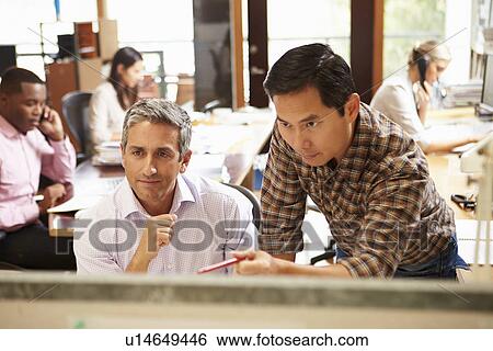 Stock Photograph - Two Colleagues Working At Desk With Meeting In Background. Fotosearch