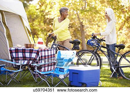 Stock Image - Two Mature Women Riding Bikes On Camping Holiday. Fotosearch