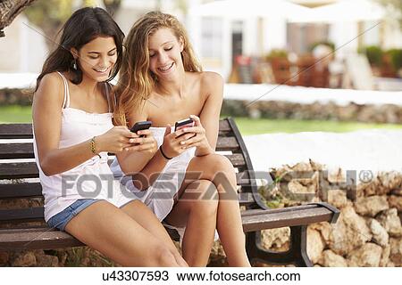 Stock Image - Two Teenage Girl Using Mobile Phone Sitting On Park Bench. Fotosearch