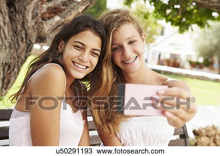 Stock Image - Two Teenage Girls Sitting On Bench Taking Selfie In Park. Fotosearch
