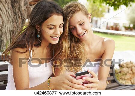 Two Teenage Girls Using Mobile Phone Sitting On Park Bench View Large Photo Image Stock Photo - Two Teenage Girls Using Mobile Phone Sitting On Park Bench. Fotosearch